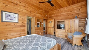 A wooden cabin interior with a bed, a television, a wicker chair, and a ceiling fan.