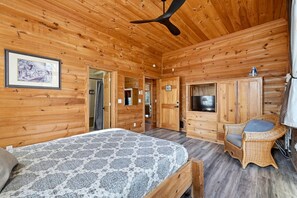 A wooden cabin interior with a bed, a television, a wicker chair, and a ceiling fan.