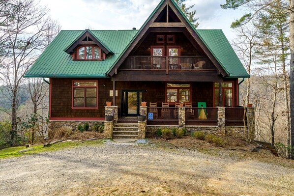 A two-story log cabin with a green metal roof, a front porch with wooden railings, and a stone foundation.
