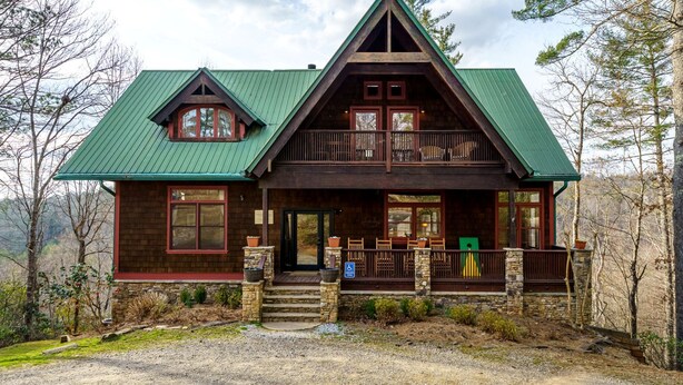 A two-story log cabin with a green metal roof, a front porch with wooden railings, and a stone foundation.