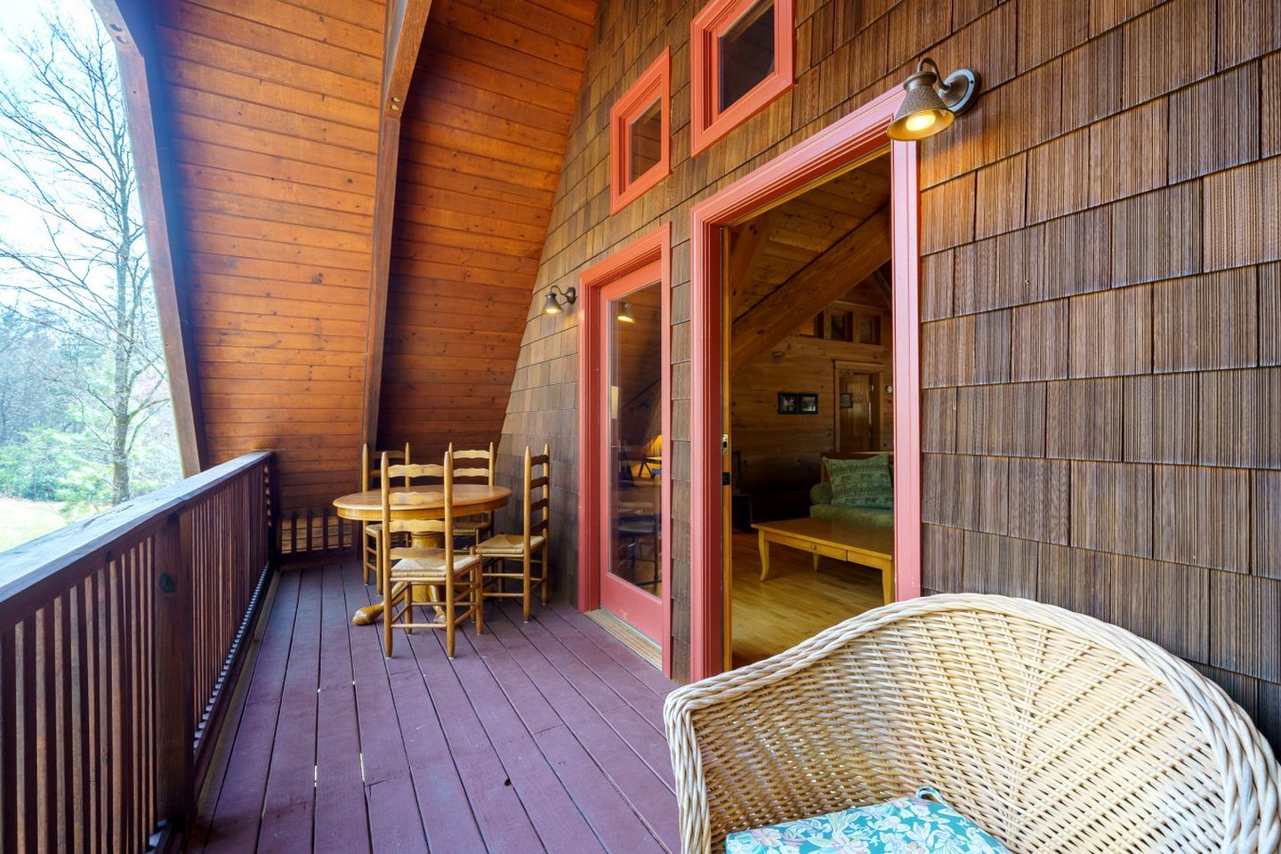 A wooden deck with a round table and chairs, a wicker chair, and a red-framed door leading to an interior space.