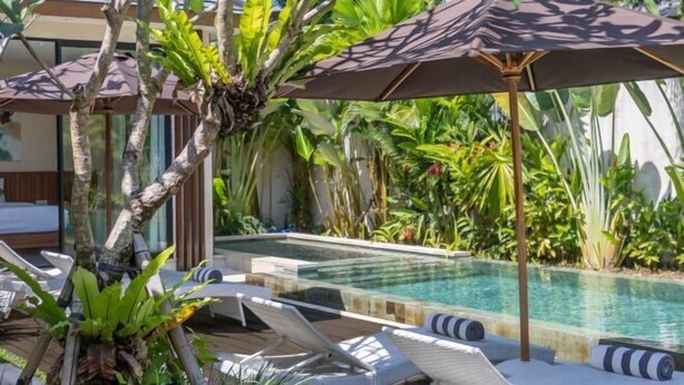 Zona junto a la piscina con sillones de descanso y sombrillas, rodeada de vegetación exuberante.