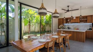 Comedor moderno con una mesa de madera dispuesta para seis personas, rodeada de lámparas colgantes de mimbre y vistas a exuberante follaje a través de amplios ventanales.