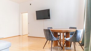 A modern living room with a wooden dining table, four grey upholstered chairs, a flat-screen TV mounted on the wall, and a light-colored wooden floor. - Basic 2BR Apt Near Westbahnhof (Vienna)