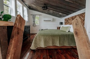 A bedroom with a wooden ceiling, a bed with a green bedspread, a dresser, a window with curtains, and a plant.