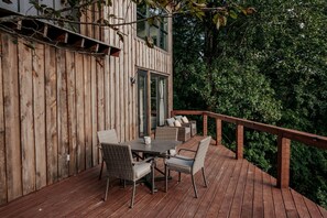 A wooden deck with a table and chairs, a glass door, and a view of a forested area.