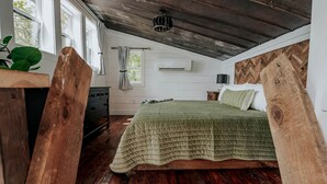 Une chambre avec un plafond en bois, un lit recouvert d’une couverture verte, une commode, une fenêtre avec des rideaux et une plante.