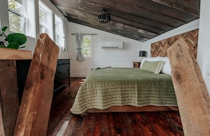 Ein Schlafzimmer mit Holzboden, einem Bett mit grüner Bettdecke, einem Schminktisch, einem Fenster mit Vorhängen und einer Pflanze.