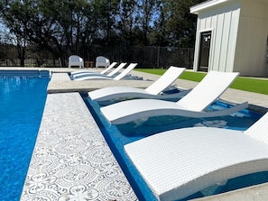 Une piscine bordée de chaises longues blanches et d’une mosaïque décorative.