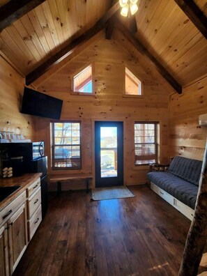 Une cabane avec un plafond mansardé, un canapé, un coin cuisine et une grande fenêtre.