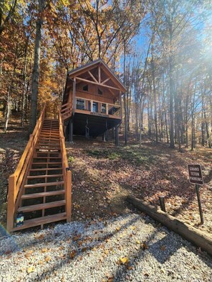 A wooden cabin with a wooden staircase leading to a deck, surrounded by trees and gravel ground.