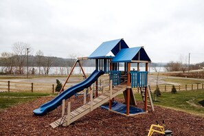 A playground with a slide and climbing structures, surrounded by a fence and a body of water in the background.