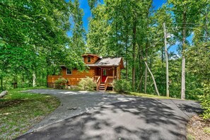 A cabin with a porch, surrounded by trees, and a paved driveway leading to it.