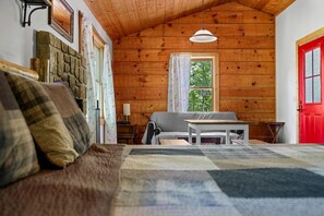 A cozy cabin interior with a wooden ceiling, a red door, a stone fireplace, a sofa, and a table.