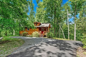 A cabin with a porch, surrounded by trees, and a paved driveway leading to it.