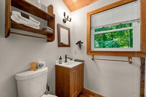 A bathroom with a toilet, sink, wooden shelves with towels, a mirror, a hairdryer, and a window with a view of greenery. - Charming Cabin with Spa and Mountain Views in Sevierville, Tennessee (Sevierville)