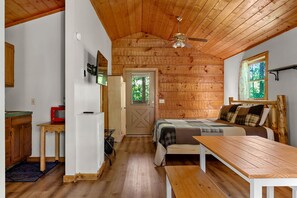 L’intérieur d’une cabane en bois, avec un lit, une table, un ventilateur de plafond et une fenêtre.