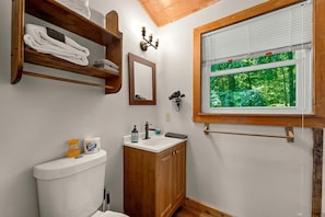 A bathroom with a toilet, sink, wooden shelves with towels, a mirror, a hairdryer, and a window with a view of greenery. - Charming Cabin with Spa and Mountain Views in Sevierville, Tennessee (Sevierville)