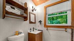 A bathroom with a toilet, sink, wooden shelves with towels, a mirror, a hairdryer, and a window with a view of greenery.