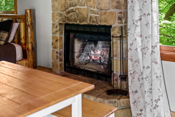 Una chimenea de piedra con fuego, una mesa de madera y un ventanal con vistas a la vegetación.