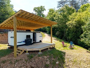 A recreational vehicle with a covered porch and a small deck, surrounded by trees and a dirt path.