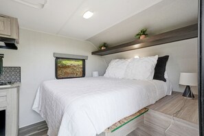 A bedroom with a bed, a nightstand, a lamp, a shelf with plants, and a window with an exit sign.