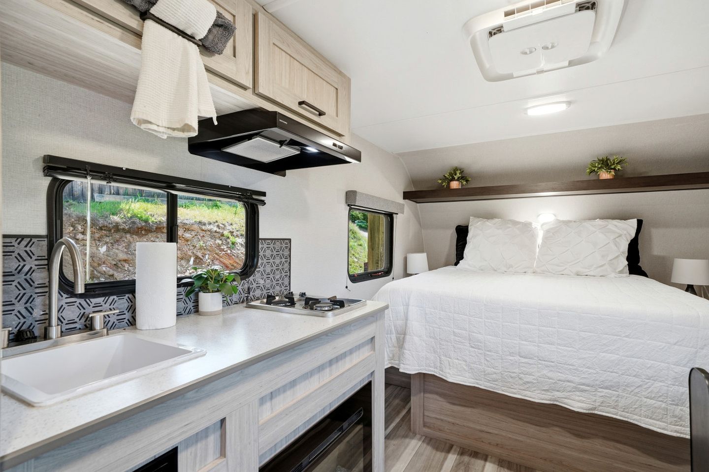 A compact RV interior with a kitchenette, a bed, and a window with a view of greenery.