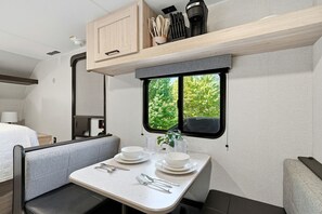 A compact RV interior with a dining area, a bed, and a window with a view of greenery.