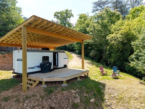 Uma casa rodante com varanda coberta e um pequeno deck, cercada por árvores e uma trilha de terra.