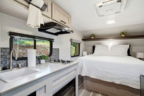 A compact RV interior with a kitchenette, a bed, and a window with a view of greenery.