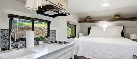 A compact RV interior with a kitchenette, a bed, and a window with a view of greenery.