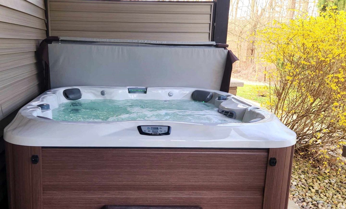 A hot tub with a control panel, surrounded by wooden walls and a garden with yellow flowers.