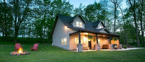 A two-story house with a covered porch, a fire pit, and two Adirondack chairs.