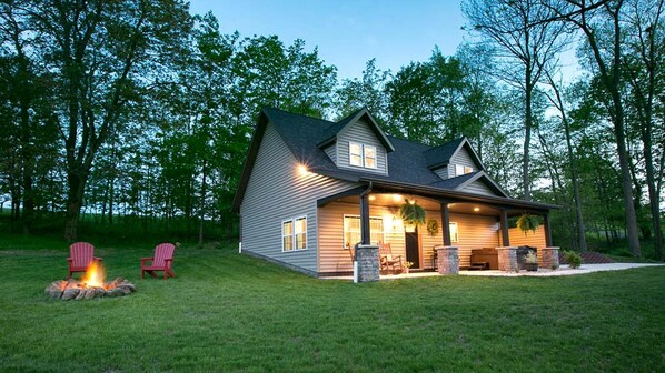 A two-story house with a covered porch, a fire pit, and two Adirondack chairs.