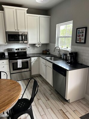 A kitchen with white cabinets, stainless steel appliances, a window, and a dining area with a wooden table and black chairs. - Cozy Cabin in the Texan Countryside for Glamping Vacations in Wimberley (Wimberley)
