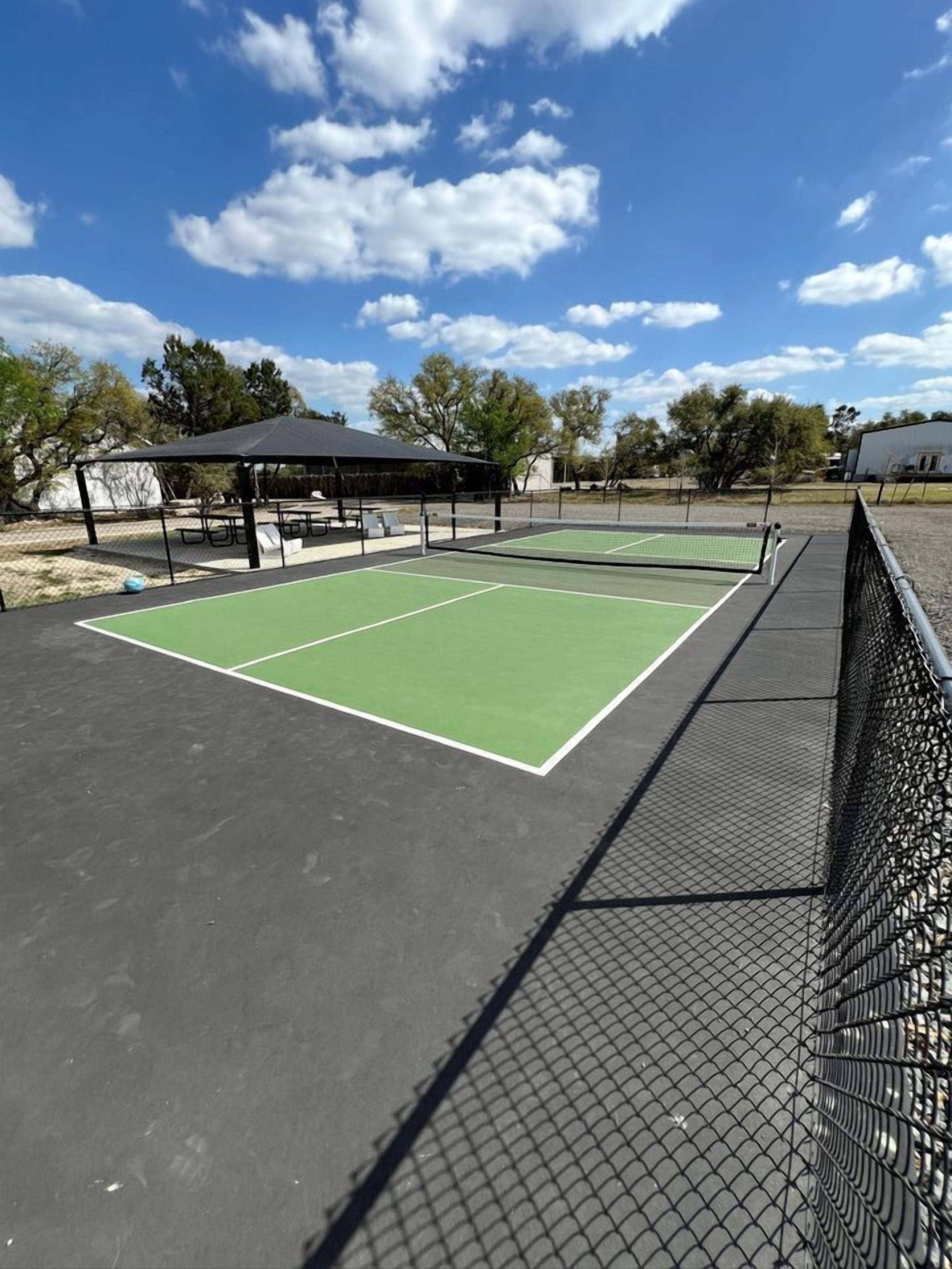 An outdoor tennis court with a net, surrounded by a fence, and a covered seating area.