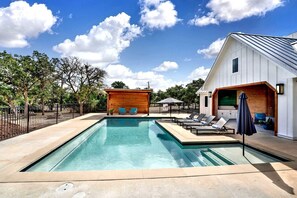 Una piscina con sillas de descanso y sombrillas, rodeada por una valla de madera y un edificio blanco con techo inclinado.