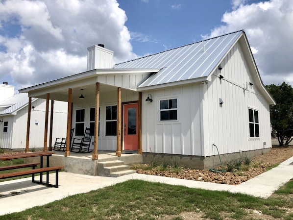 Una casa blanca de un solo piso con un porche cubierto, una puerta roja y un techo de metal.