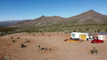 Paisaje desértico con una casa rodante, un coche rojo y un cobertizo pequeño.