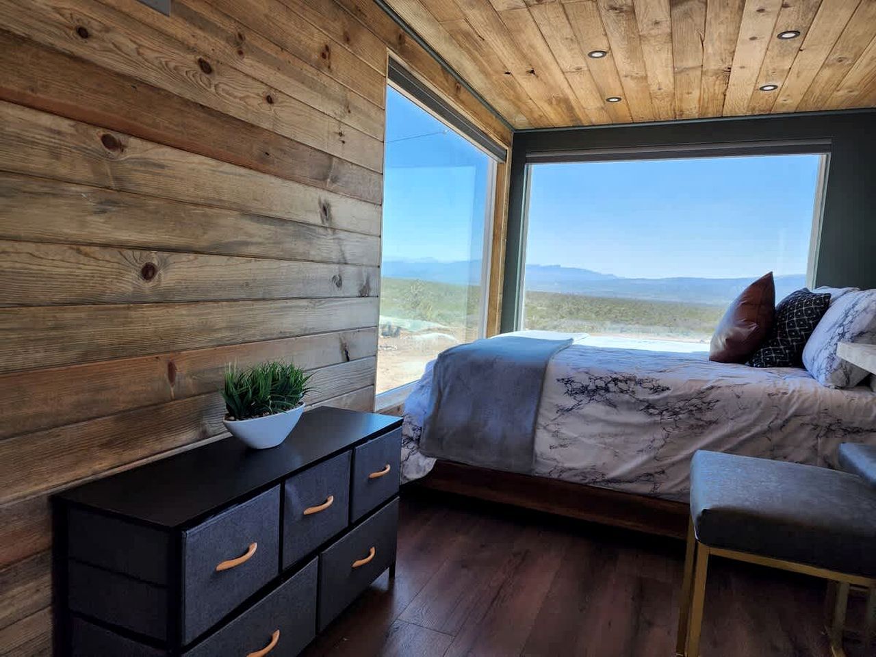 A bedroom with a wooden wall, a bed with a view, a nightstand, and a window.