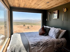 A bedroom with a large window, a bed with patterned bedding, and a desert view.