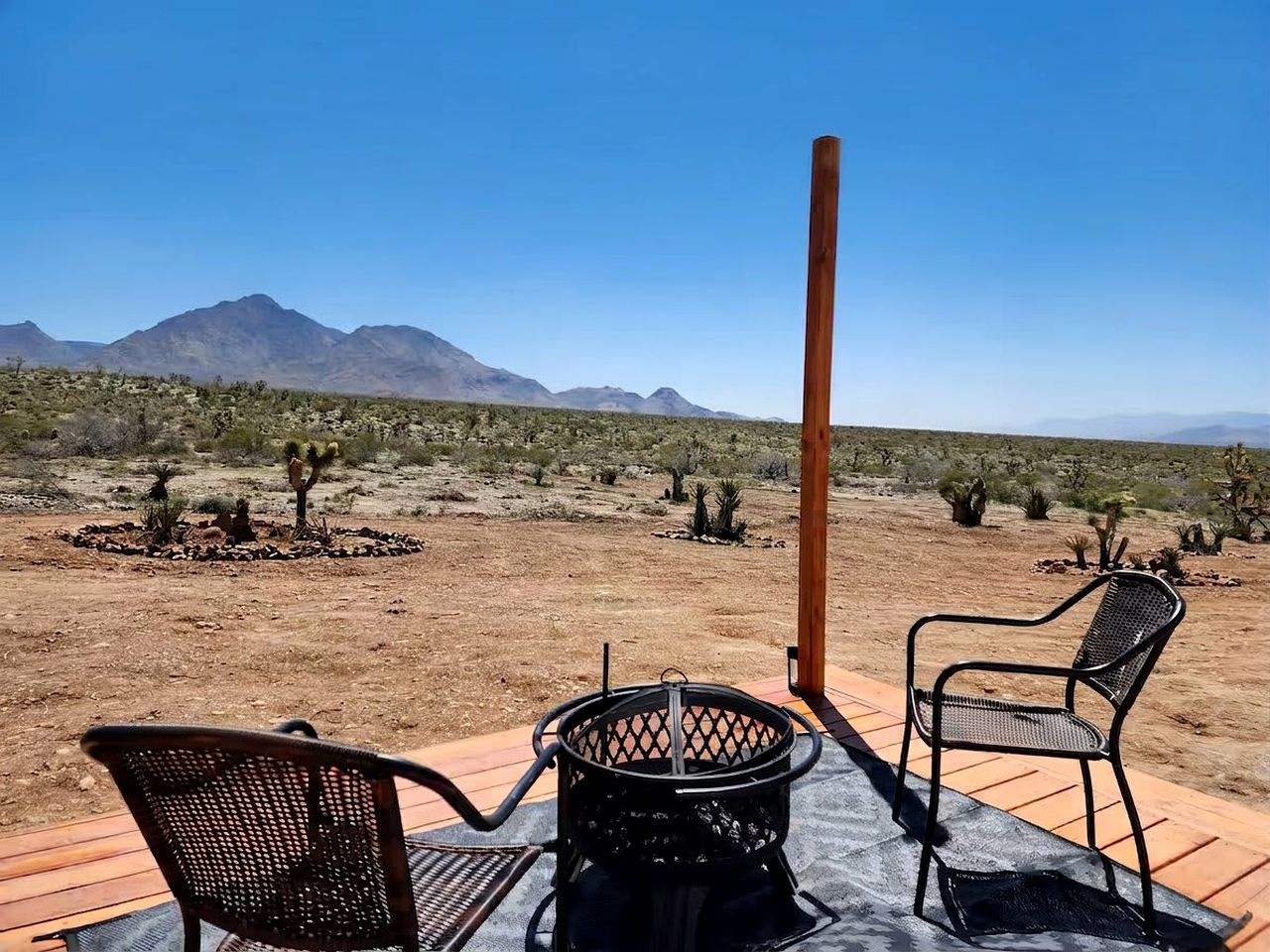 An outdoor seating area with two chairs and a metal table, overlooking a desert landscape with sparse vegetation and distant mountains.