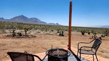 An outdoor seating area with two chairs and a metal table, overlooking a desert landscape with sparse vegetation and distant mountains.