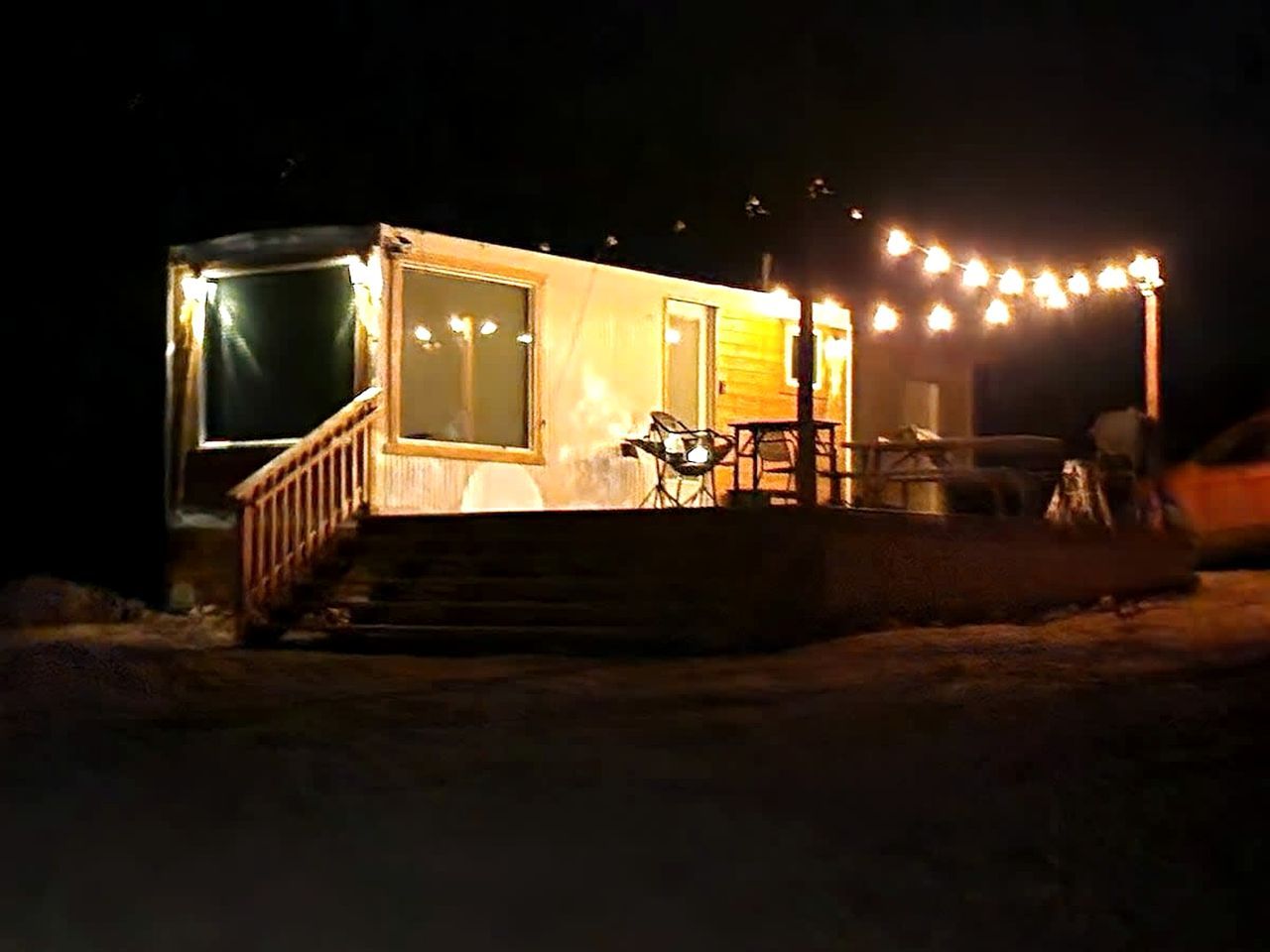 A small, illuminated cabin with a wooden deck and a single chair outside.