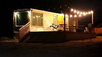 A small, illuminated cabin with a wooden deck and a single chair outside.