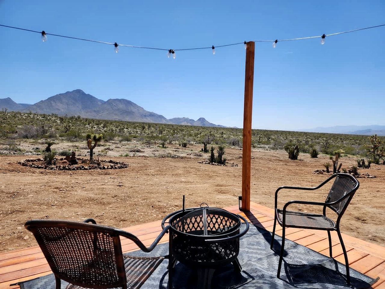 Un patio al aire libre con dos sillas y una mesa, un hogar de fuego metálico y luces colgantes frente a un paisaje desértico con montañas al fondo.