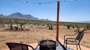 Un patio al aire libre con dos sillas y una mesa, un hogar de fuego metálico y luces colgantes frente a un paisaje desértico con montañas al fondo.