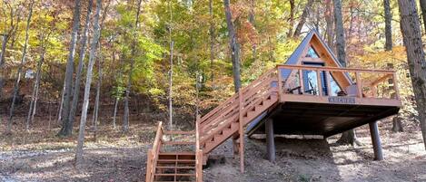 A wooden cabin with a staircase leading to a deck, surrounded by trees in autumn colors.