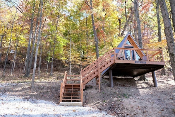 A wooden cabin with a staircase leading to a deck, surrounded by trees in autumn colors.