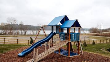 A playground with a slide and climbing structures, surrounded by a fence and a body of water in the background.